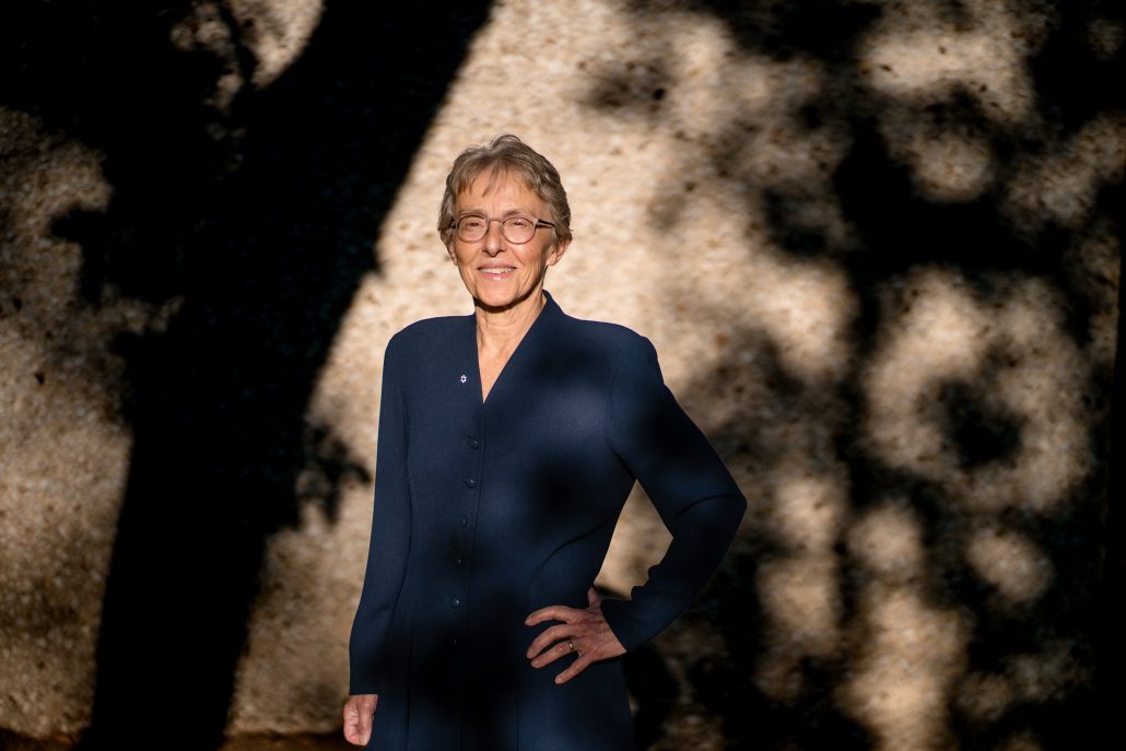 Woman standing in the sun with shadows from surrounding trees on the wall behind her.