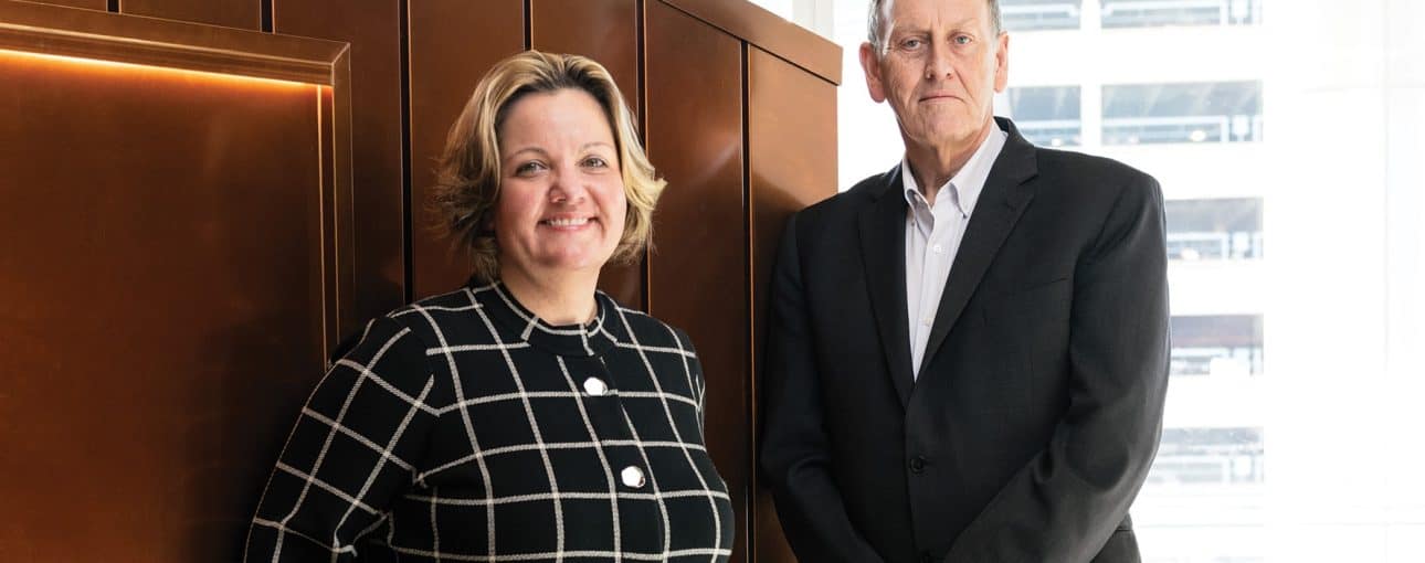 Man and woman in business attire leaning against a copper-coloured wall.