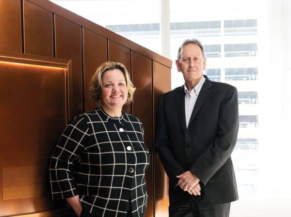 Man and woman in business attire leaning against a copper-coloured wall.