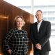 Man and woman in business attire leaning against a copper-coloured wall.