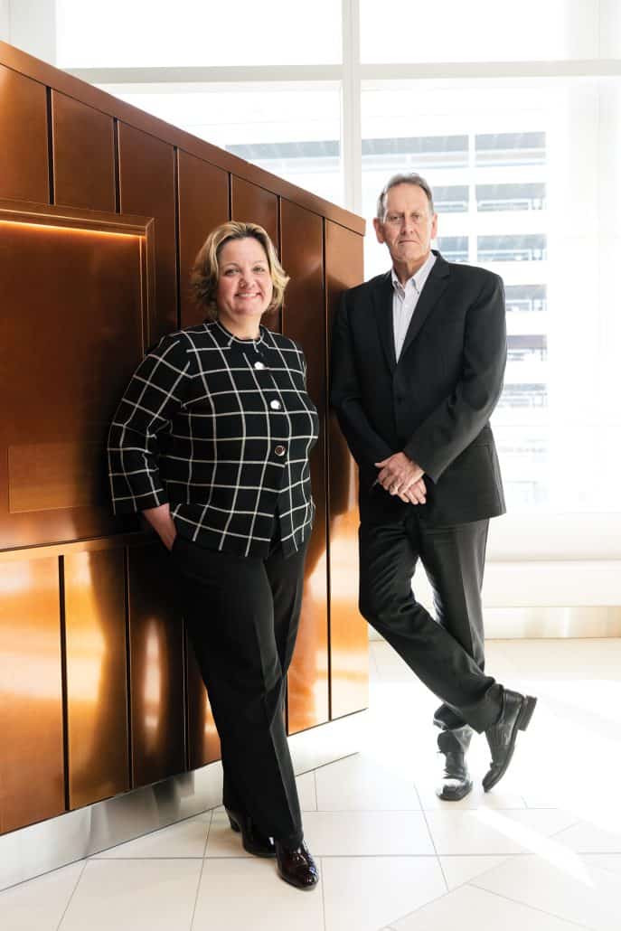 Arthur J.E. Child Comprehensive Cancer Centre 1 Amanda Weiss and Dr. Don Morris in business wear standing up against a copper-coloured wall.