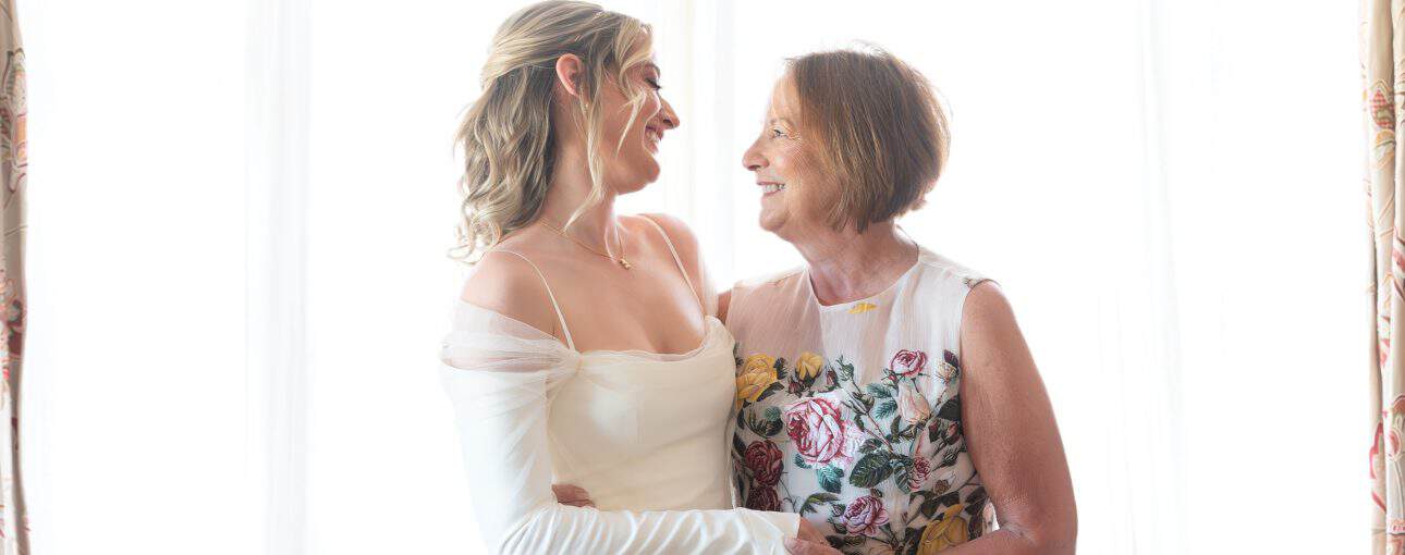Mother and daughter hugging each other. The daughter is wearing a white wedding dress and mom in is a white dress covered in colourful flowers.