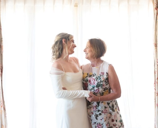 Mother and daughter hugging each other. The daughter is wearing a white wedding dress and mom in is a white dress covered in colourful flowers.