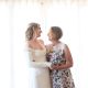 Mother and daughter hugging each other. The daughter is wearing a white wedding dress and mom in is a white dress covered in colourful flowers.
