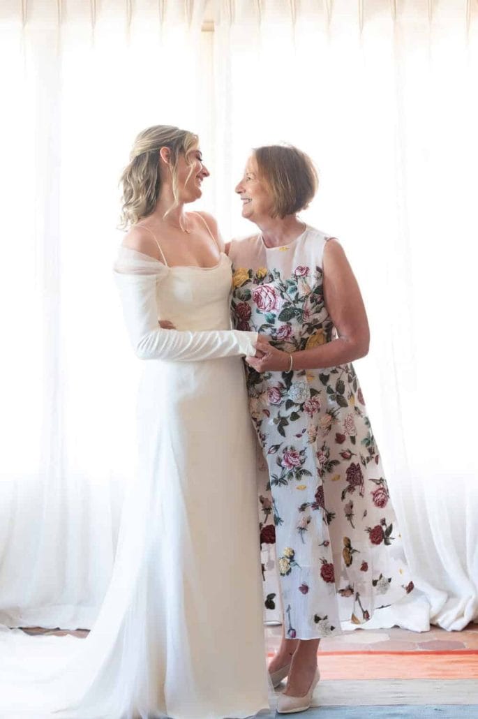 More time, across generations 1 Andrea and Alexandra McManus hugging each other. Alexandra is wearing a white wedding dress and Andrea in is a white dress covered in colourful flowers.