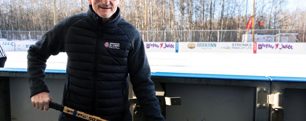 Barrie Stafford stands in front of an ice rink, holding a hockey stick.