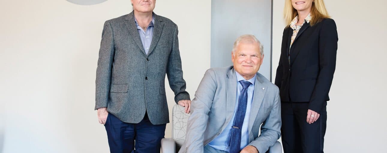 A picture of three medical professionals. Two stand while the third sits in a chair between them.