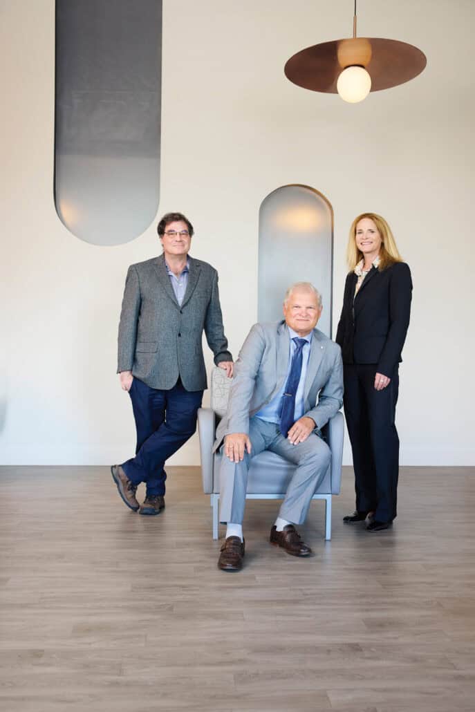 A picture of three medical professionals. Two stand while the third sits in a chair between them.
