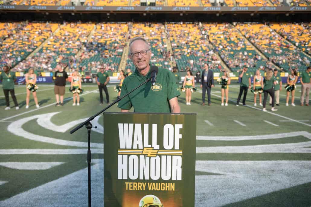 A voice of hope 1 Man stands in front of a podium with a professional football field, stands and cheerleaders behind him.