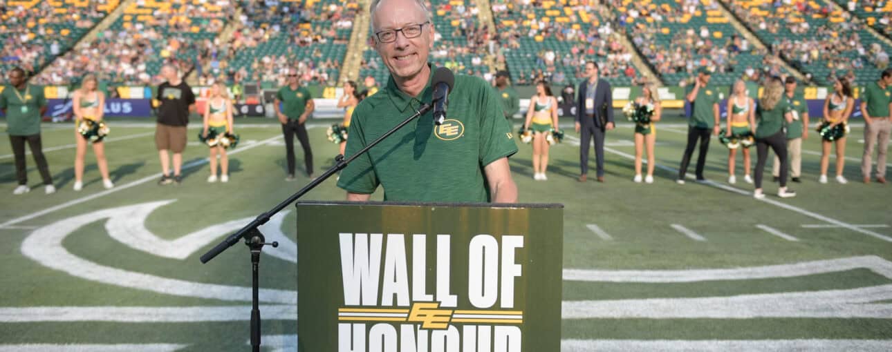 Man stands in front of a podium with a professional football field, stands and cheerleaders behind him.