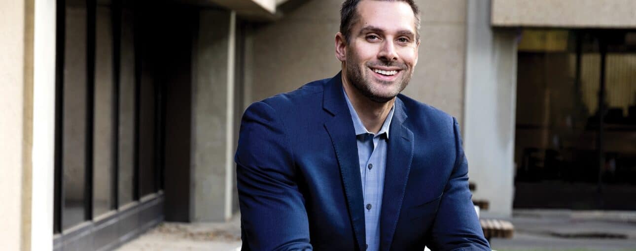 A man in a blue suit jacket sits on a park table with his legs up on the bench, smiling right at the camera.