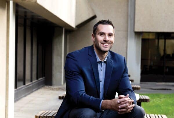 A man in a blue suit jacket sits on a park table with his legs up on the bench, smiling right at the camera.