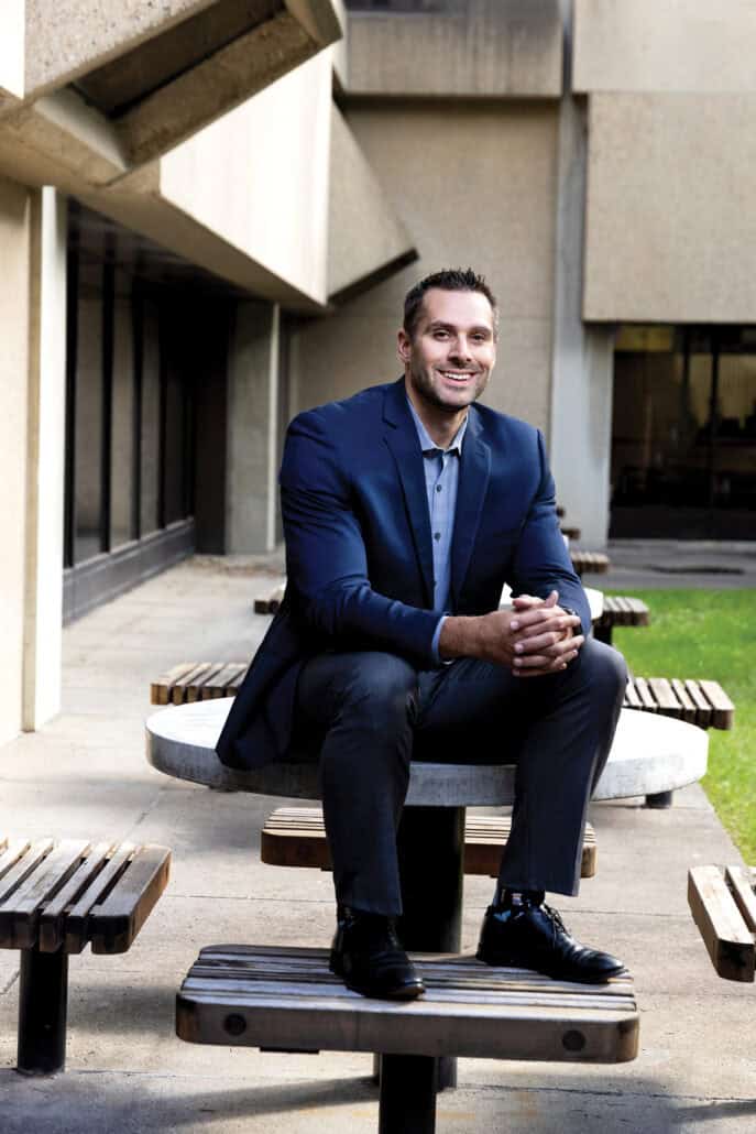 A man in a blue suit jacket sits on a park table with his legs up on the bench, smiling right at the camera.