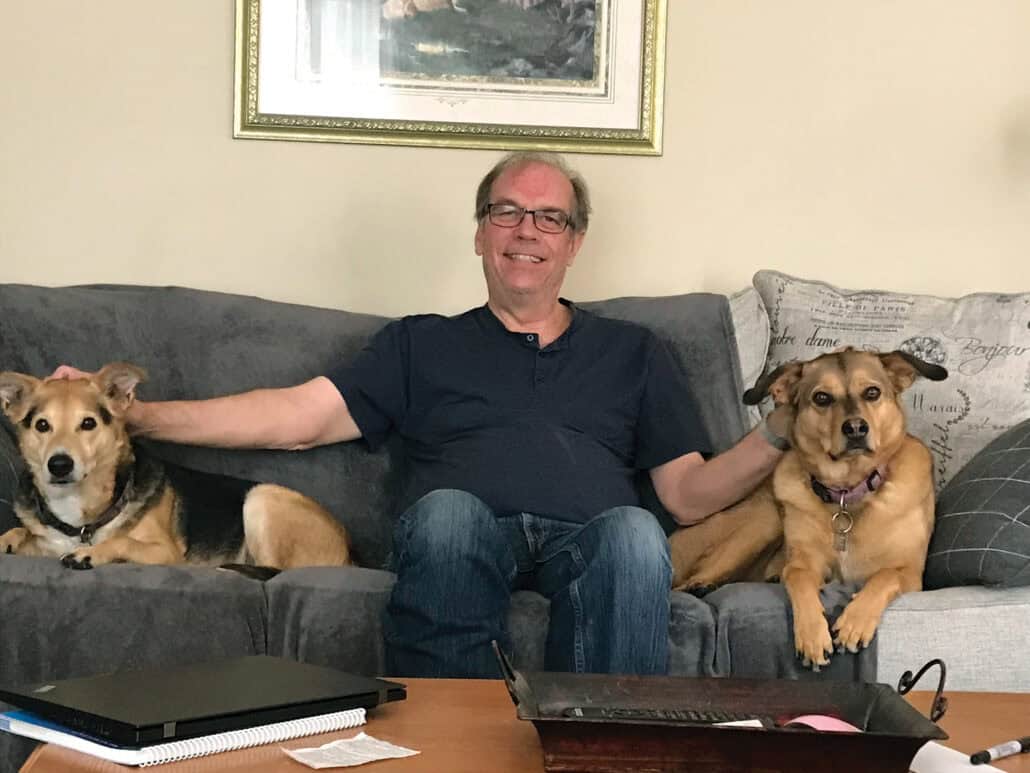 A man sits on the couch while petting two dogs, one on each side of him.