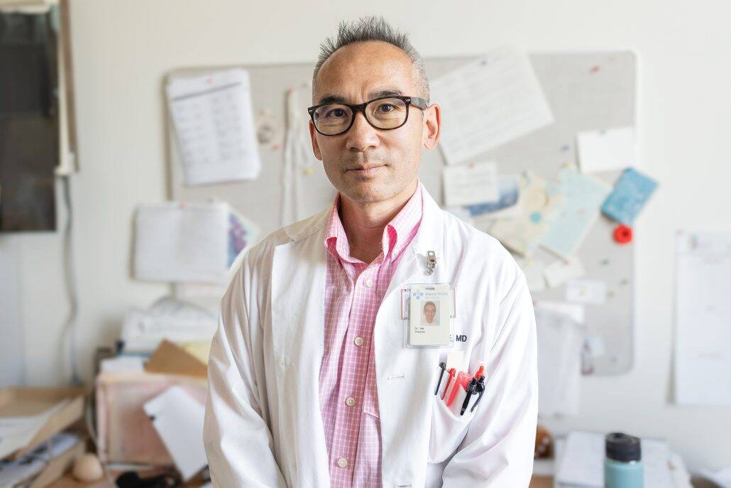 Leading the way. 3 Man in a lab coat and pink shirt looking at the camera with papers on a bulletin board behind him.