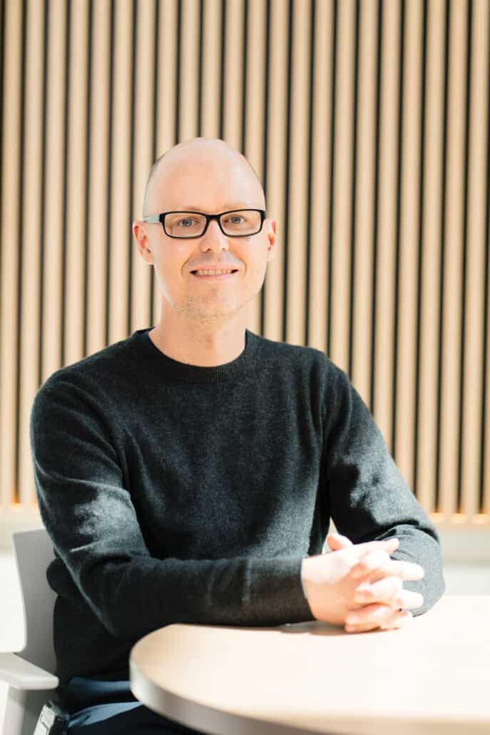 Dr. Matthew Anaka sits in front of a wood-panelled wall with his fingers interlaced.