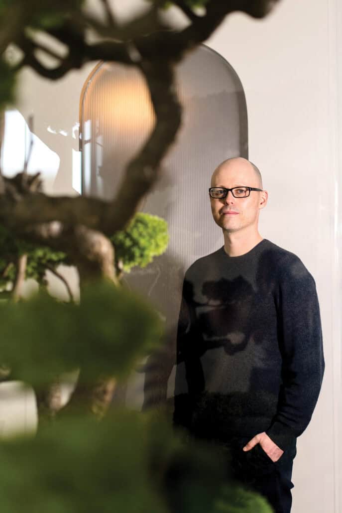 Dr. Matthew Anaka stands, with his hand in his pocket, in the rear of the image, with part of a plant in the foreground. 