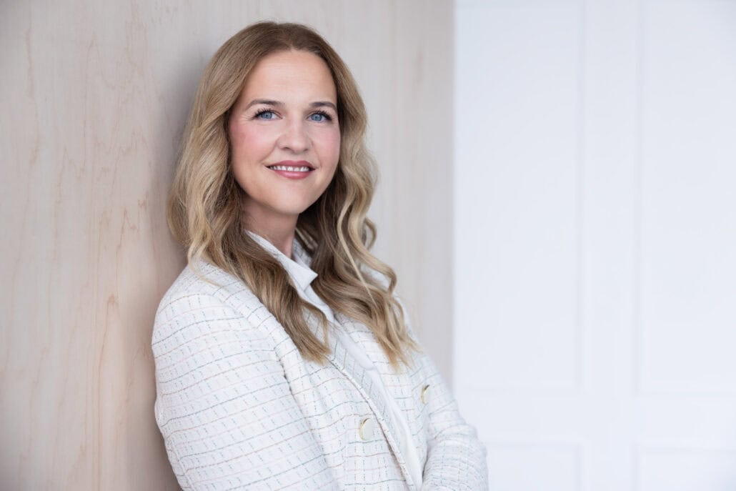 Woman in a white jacket leans against a wall and smiles at the camera.