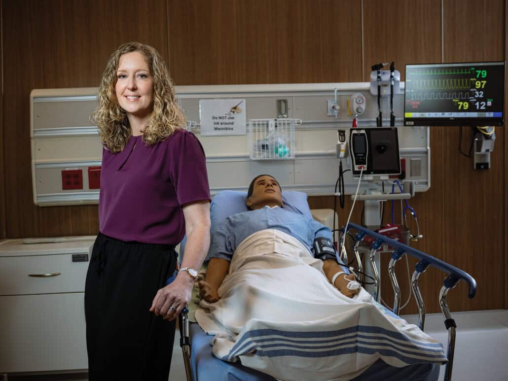 Jillian Gautschi-Nichol stands next to a pretend patient (manikin) in a hospital bed.