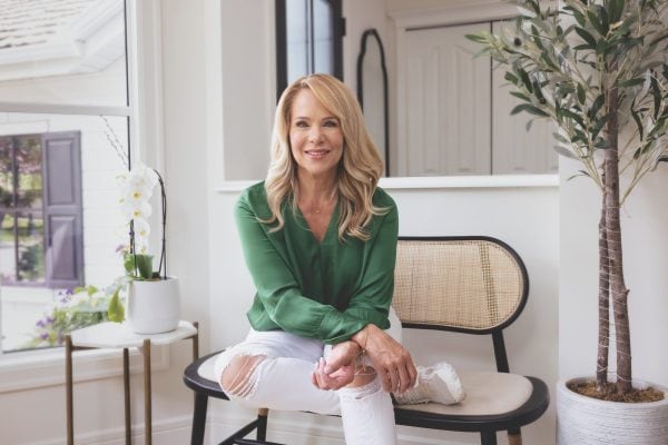 Woman in a green shirt and white pants sitting on a bench near the entryway of a home with an indoor plant next to her.