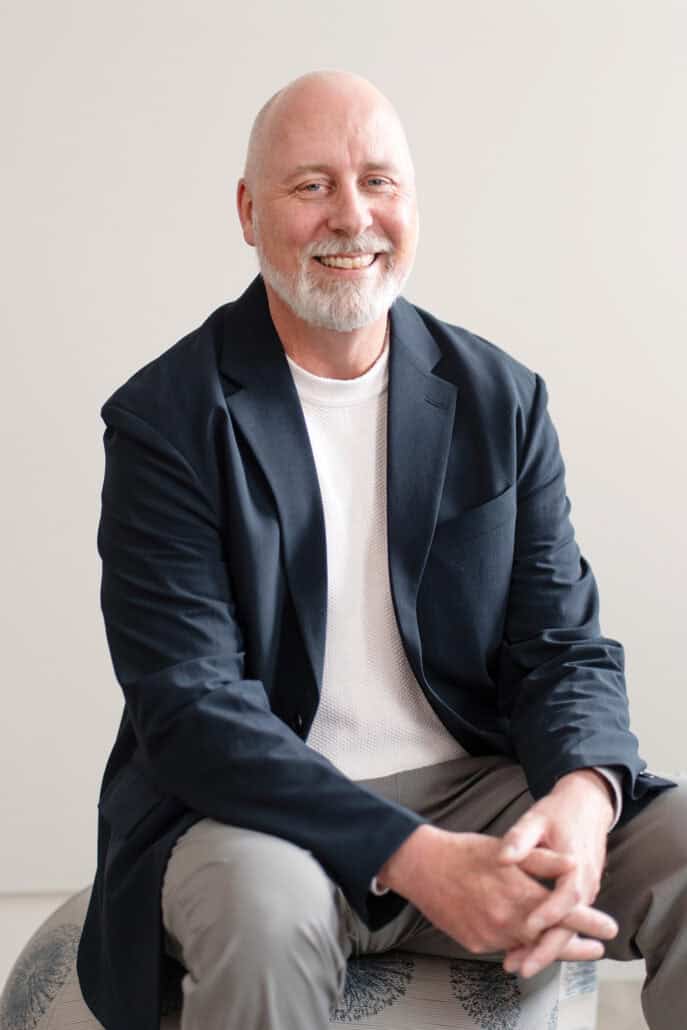 Portrait of Dr. John Walker, sitting in a navy blazer and with his finger interlaced in his lap.