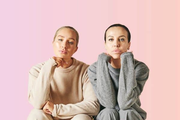 Two women pose looking into the camera with a pink background.