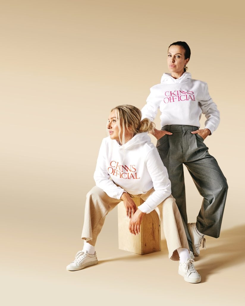 Two women pose in white sweatshirts with a beige backdrop. 