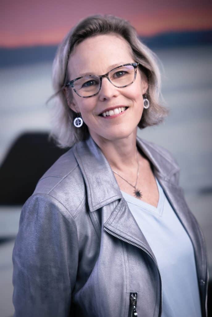 Dr. Linda Carlson wearing  a silver jacket and glasses looks into the camera and smiles.