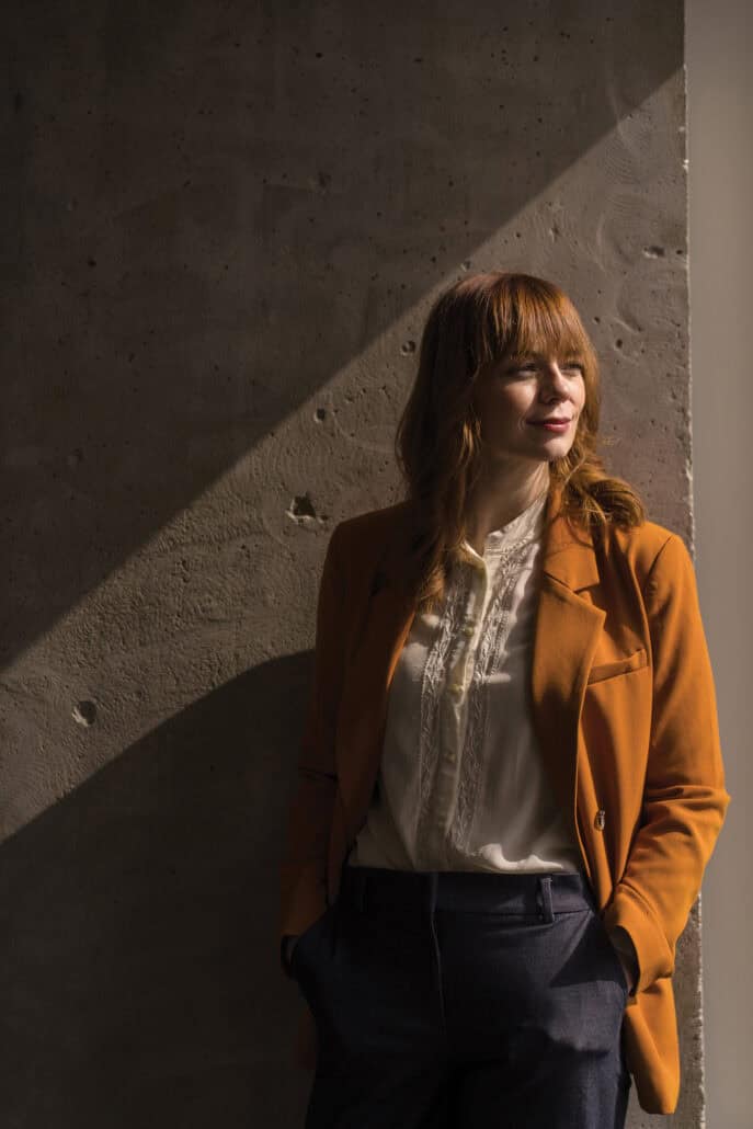 Maeve Wickham in a burnt orange blazer, standing against a wall, her hands in her pocket and looking off to the side, right some shadows behind her. 