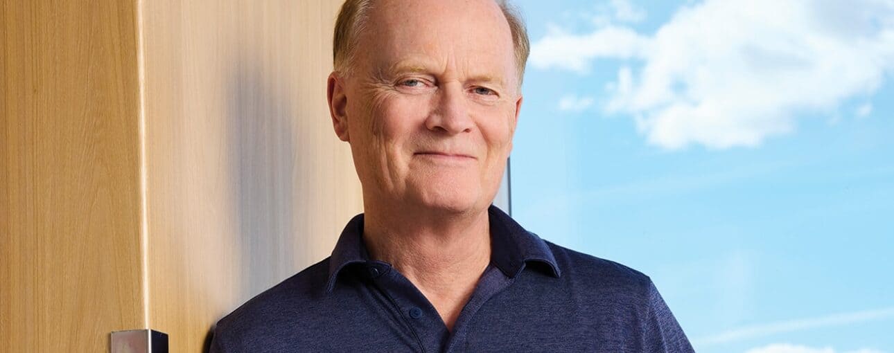 Man in a blue shirt leans against a wall with a window and blue skies behind him.