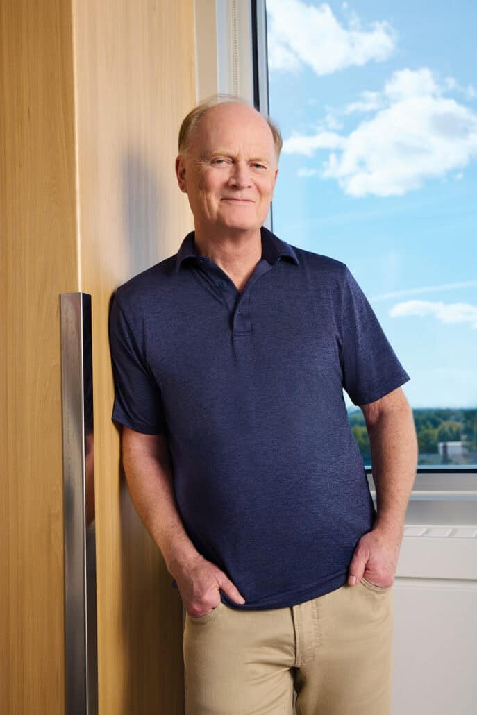 Man in a blue shirt leans against a wall with a window and blue skies behind him.