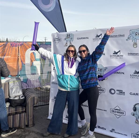 Two women smile with their arms raised in front of the Cold Patrol 10K sponsorship board.