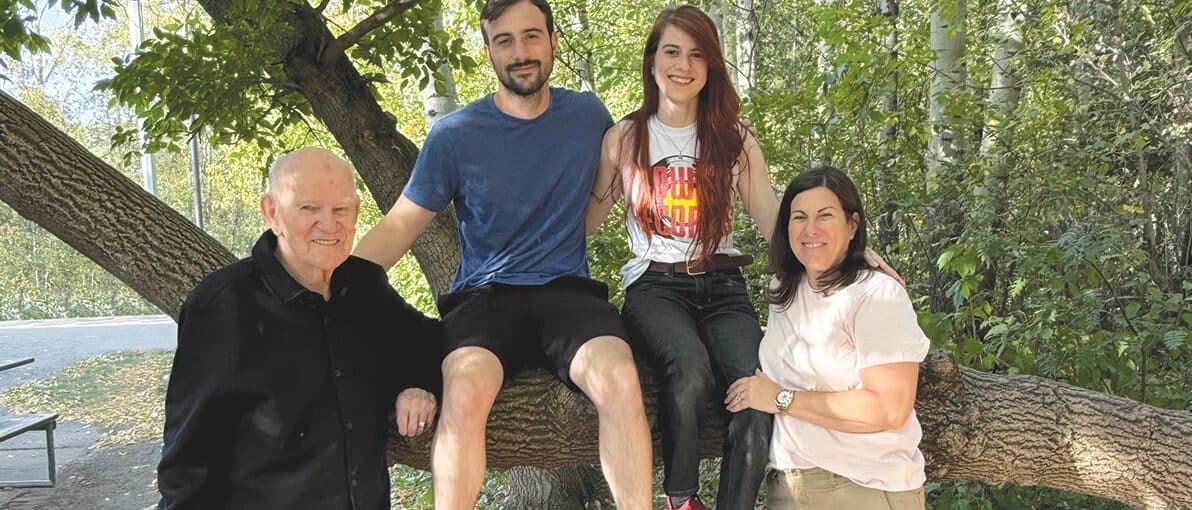 Peter Kossowan stands next to a large tree branch which his grandson and granddaughter are sitting on, along with his daughter on the other side.