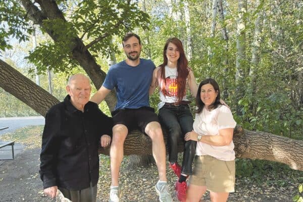 Peter Kossowan stands next to a large tree branch which his grandson and granddaughter are sitting on, along with his daughter on the other side.