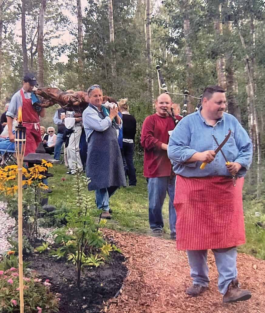 Gifts fuelled by love 3 Men walk on a trail in the woods while carrying a spit-roast pig.
