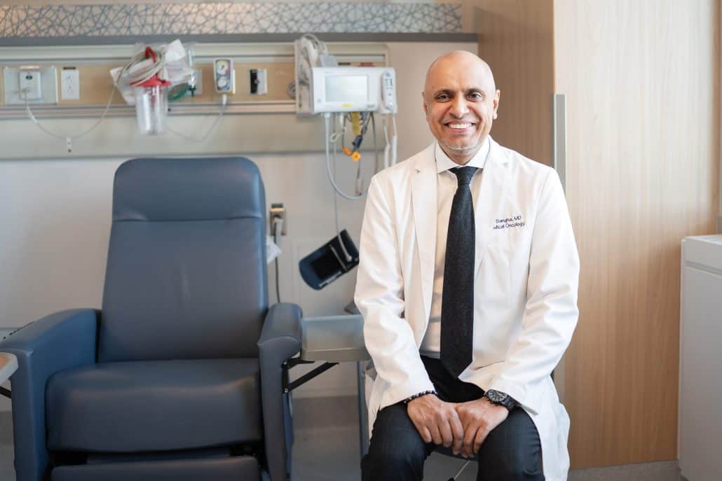 Doctor in a white coat smiles at the camera as he sits next to a patient chair.