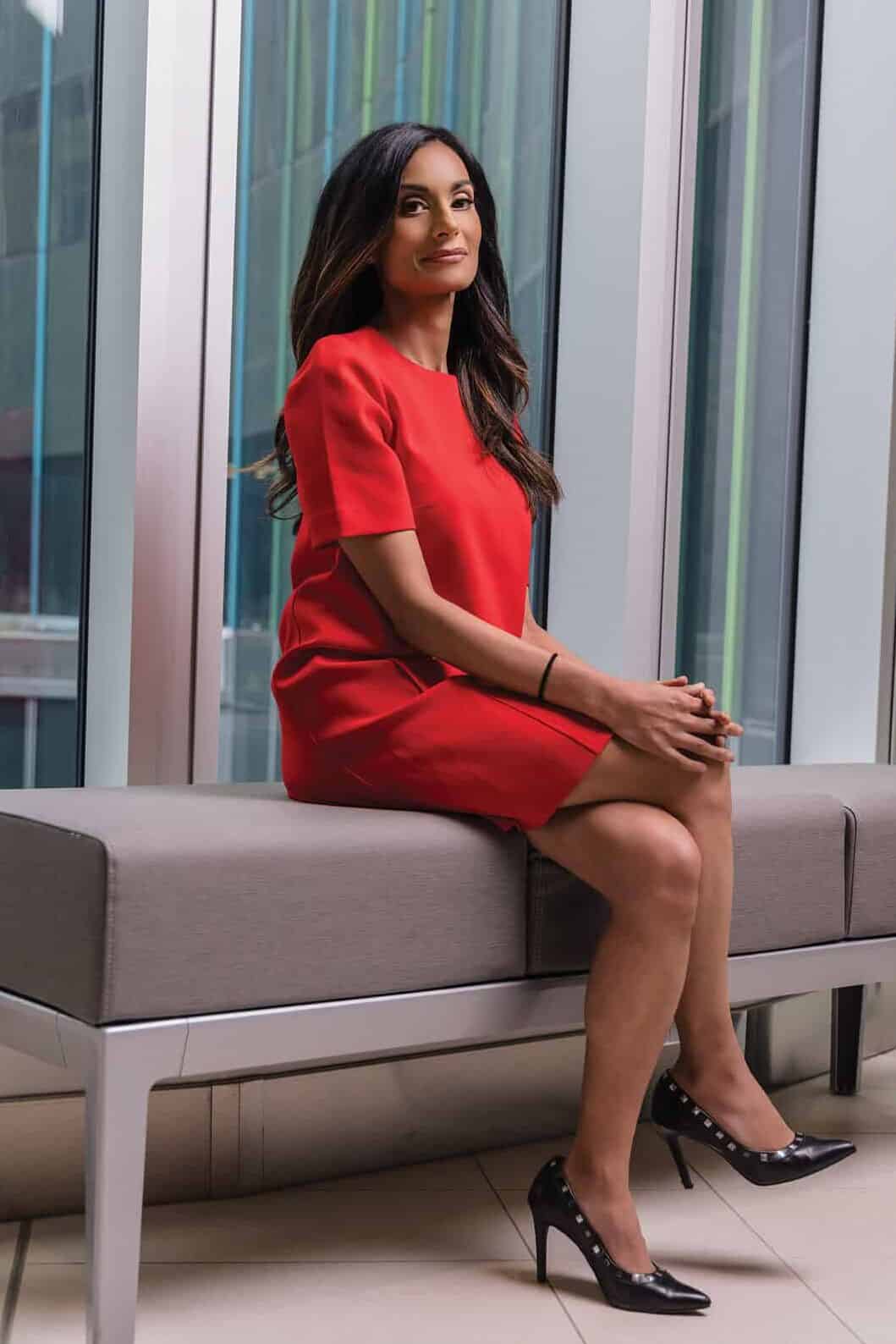 Dr. Safiya Karim sits with her legs crossed, wearing a bright red dress and heels.