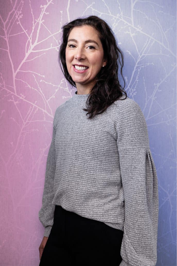 Woman in a grey sweater stands against a blue and purple wall. 