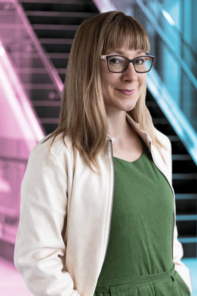 Female doctor in white lab coat and green shirt with a pink and blue background.