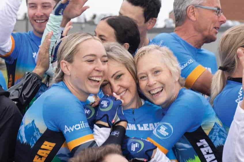 Sydney Meier and Leah Meier hold each other, smiling in celebration, alongside their cycling teammates.