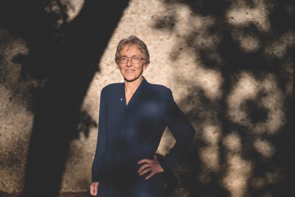 Woman standing in the sun with shadows from surrounding trees on the wall behind her.